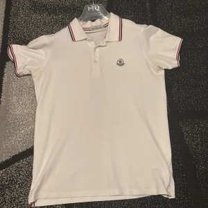 Vit pikétröja från Moncler M - Snygg vit pikétröja från Moncler i storlek M. Klassisk design med Moncler-logga på bröstet och krage med röda och marinblå ränder. Tröjan har korta ärmar och knäppning framtill. Perfekt för en clean och sportig look.