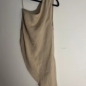 KAALA Oneshoulder Linne - Snygg beige oneshoulder topp från Bik Bok i storlek M. Klänningen har asymmetrisk axel, smal passform och dragkedja i sidan. Perfekt för dig som gillar minimalistisk stil och vill sticka ut med en enkel men trendig look. Ny med prislapp 