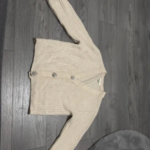 Beige ribbad kofta med glittriga knappar - Superfin beige kofta med ribbat mönster och långa ärmar. Koftan har v-ringning och fyra stora, glittriga knappar som verkligen poppar. Perfekt för dig som gillar en mysig men ändå stylish look. Materialet känns mjukt och lite fluffigt.