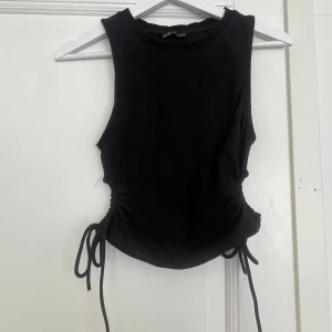 Svart ribbad linnetopp från Zara - Säljer en svart ribbad linnetopp från Zara med snygga knytband i sidorna. Toppen är ärmlös och har en croppad passform som ger en cool och trendig look. Perfekt att matcha med höga jeans eller kjol för en stilren outfit.