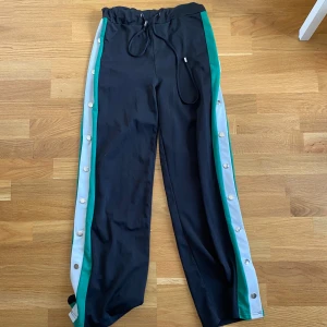 Svarta byxor med knappar - Snygga svarta byxor gröna och vita ränder längs sidorna. Byxorna har snörning i midjan och tryckknappar längs benen för en sportig vibe. Perfekta för chill.