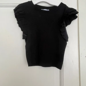 Svart volangtopp från Zara - Supersnygg svart topp från Zara med volangdetaljer på axlarna. Modellen är ärmlös och har en tight passform som framhäver siluetten. Perfekt att styla med jeans eller kjol för en trendig look.
