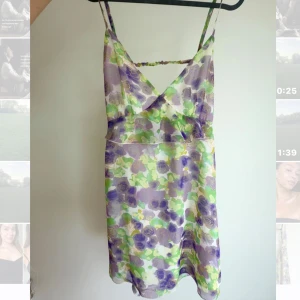 Blommig kort klänning med öppen rygg - Superfin kort klänning med lila och grönt blommönster, tunna axelband och v-ringning. Klänningen har öppen rygg med snygg detalj och är gjord i ett lätt, glansigt material som känns som polyester eller satin. 