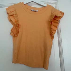 Ribbad volangtopp från Zara i orange - Superfin ribbad topp från Zara i en ljus orange färg. Toppen har ärmlös design med breda volanger på axlarna som ger en extra snygg touch. Perfekt för dig som gillar att sticka ut lite extra med detaljer.