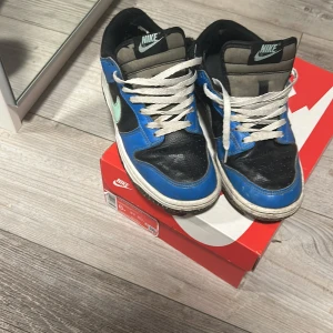 Nike Dunk Low blå och svarta sneakers - Nike Dunk Low sneakers i svart och blått med vit sula och vit snörning. Swoosh-loggan är mintgrön och skorna har en klassisk låg siluett. Ovandelen är i skinn och textil, med detaljer i grått på plösen. Perfekta för dig som gillar streetwear och retrostil.