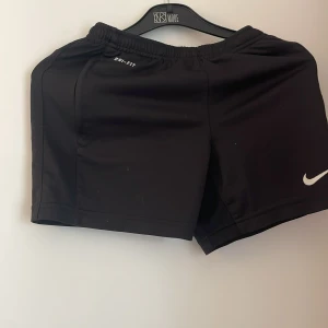 Svarta Nike Dri-Fit träningsshorts - Svarta träningsshorts från Nike med Dri-Fit-teknologi och vit logga på benet. De har elastisk midja och är perfekta för sport eller gym. Snygg och enkel design som funkar till alla träningspass.