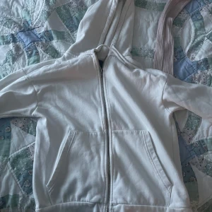 Vit hoodie med dragkedja och huva - Klassisk vit hoodie med dragkedja framtill och två stora fickor. Tröjan har huva och långa ärmar, perfekt för en chill och avslappnad stil. Gjord i mjukt bomullsmaterial som känns skönt mot huden.