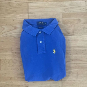 Ralph Lauren piké  - Klassisk blå pikétröja från Ralph Lauren med gul broderad logga på bröstet. Perfekt för en clean look. Storlek S skulle även passa folk som bär M