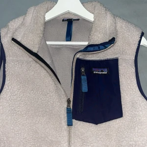 Beige fleeceväst från Patagonia - Säljer en beige fleeceväst från Patagonia med marinblå detaljer och dragkedjor. Västen har en hög krage, tre fickor med blå dragkedjor och en snygg bröstficka med logga. Perfekt lager-på-lager-plagg för kyliga dagar. Använd ett fåtal gånger, inte än ända fläck överhuvudtaget, as skön att gå runt med, säljer då den är för liten!