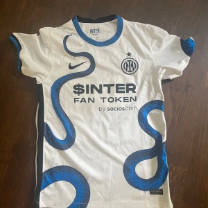 Inter Milan bortatröja  - Inter Milans officiella bortatröja med Barella 23 på ryggen. Vit bas med blå ormmönster som slingrar sig över tröjan, svarta detaljer och blå krage. Nike-logga och klubbmärke på bröstet, tillverkad i lätt funktionsmaterial. Snygg och unik design för dig som älskar fotboll och Inter.