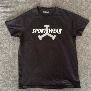 Svart sportig t-shirt från Tridri - Svart tränings t-shirt från Tridri med trycket 'SPORTSWEAR' och en stor logga på bröstet. Klassisk rund hals och korta ärmar. Tillverkad i en mjuk mix av polyester och viskos som känns lätt och skön mot huden. Perfekt för dig som gillar en sportig vibe.