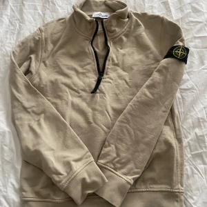 Beige half zip tröja Stone Island Junior 8 år - Snygg beige långärmad tröja från Stone Island med svart half zip-dragkedja och klassisk patch på ärmen. Tröjan har hög krage och är gjord i mjuk bomull, perfekt för lager-på-lager och chill stil. Tröjan är i mycket gott skick och endast lite skrynklig på bilderna. 