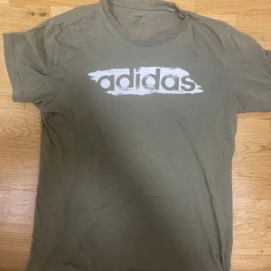Olivgrön Adidas t-shirt med tryck - Olivgrön t-shirt från Adidas med stort vitt logotryck över bröstet. Klassisk rund halsringning och korta ärmar. Skön och avslappnad passform, perfekt till jeans eller shorts. Tillverkad i mjuk bomull för en bekväm känsla hela dagen.