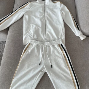 Vita mjukisbyxor från BOSS med revärer - Snygga vita mjukisbyxor från BOSS med präglad logga över hela tyget. Byxorna har elastisk midja med snörning och revärer i svart och beige längs sidorna. Perfekta för en clean och sportig look.