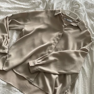 Beige skjorta från Zara i XS - Snygg beige skjorta från Zara i storlek XS. Skjortan har klassisk krage, dolda knappar framtill och långa ärmar med manschett. Materialet är mjukt och har ett lätt glansigt fall som ger en clean och stilren look. Perfekt att styla med jeans eller kjol.