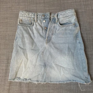 Ljusblå jeanskjol med rå kant - Snygg ljusblå jeanskjol med hög midja och rå, fransig kant nertill. Klassisk femficksmodell med knappgylf och bälteshällor. Perfekt till sneakers eller boots för en chill look.