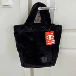 Ny svart pälsig handväska från Champion - Säljer en oanvänd svart fluffig handväska från Champion med mjuk fuskpäls och korta handtag. Väskan har innerfoder, dragkedjefack och tydlig Champion-logga framtill. Perfekt accessoar för dig som vill sticka ut med en mysig och trendig look.