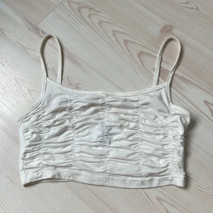 Vit ruched linnetopp från SHEIN - Säljer en vit croppad linnetopp från SHEIN i storlek S. Toppen har tunna axelband och snygga rynkade detaljer framtill med vertikala sömmar som ger en cool effekt. Perfekt till jeans eller kjol för en trendig look.