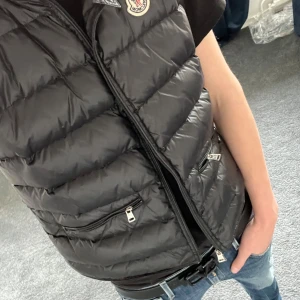 Svart dunväst från Moncler - Snygg svart dunväst från Moncler med dragkedja och hög krage. Västen har en quiltad design och två praktiska fickor med dragkedja. Perfekt till våren. Köpt på Plick och är därför lite osäker på äktheten. Dragkedjan strular lite. Storlek 2, motsvarar s-m. 