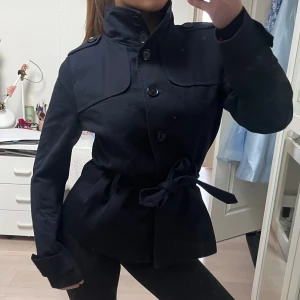 trenchcoat - Snygg svart kappa med hög krage och knappar framtill. Jackan har ett knytbälte i midjan för en elegant passform. Perfekt för kyligare dagar.