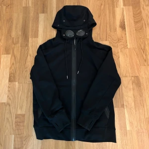 Svart jacka med huva från C.P. Company - Säljer en svart jacka från C.P. Company med unik design. Jackan har en huva med inbyggda glasögon och dragkedja framtill. Perfekt för en stilren look. Vid frågor va inte rädd att kontakta! Priset är inte hugget i sten!