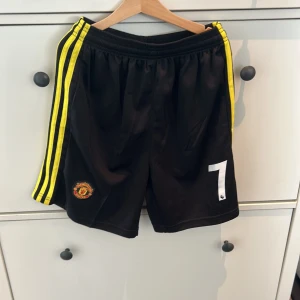Svarta Manchester United shorts med gula ränder - Snygga svarta shorts med gula ränder och Manchester Uniteds emblem. Nummer 7 tryckt på benet. Perfekta för fotbollsfans!