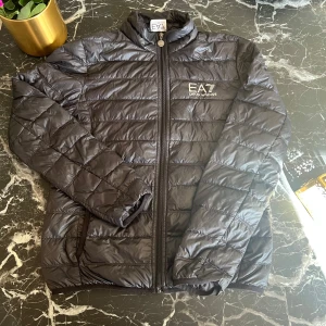 Svart pufferjacka från Emporio Armani - Snygg svart pufferjacka från Emporio Armani med EA7-logga på bröstet. Jackan har en dragkedja framtill och är quiltad för extra värme. Perfekt för kyligare dagar.