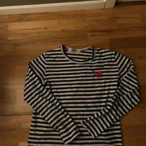 Randig långärmad tröja från Comme des Garçons - Säljer en snygg randig tröja från Comme des Garçons med det ikoniska röda hjärtat på bröstet. Tröjan är blå och vit med långa ärmar, perfekt för en stilren look. Passar både till jeans och kjol! Storleken är L men har blivit urtvättad till en storlek S ungefär