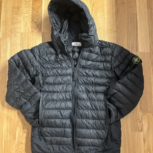 Svart dunjacka från Stone Island - Säljer en svart dunjacka från Stone Island med huva och dragkedja. Jackan har ett quiltat mönster och en ikonisk logga på ärmen. Perfekt för kyligare dagar. Har ett litet hål på höger armbåge därav priset.