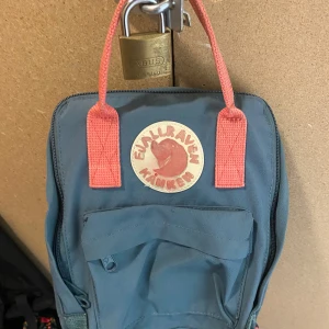 Blå ryggsäck från Fjällräven - Säljer en klassisk blå Fjällräven Kånken ryggsäck med rosa handtag och axelremmar. Ryggsäcken har en dragkedja framtill och ett stort huvudfack. Perfekt för vardagsbruk eller utflykter. En tidlös design som aldrig går ur stil.