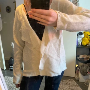 Beige/vit kofta med knappar - Säljer en stilren kofta med dekorativa knappar framtill. Koftan har långa ärmar och ett unikt mönster med korsstygn på ryggen. Perfekt för en avslappnad look.