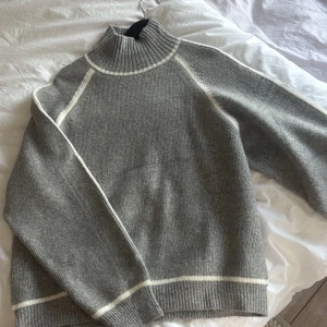 Grå stickad ull tröja från Mango - Säljer en stilren grå stickad polotröja från Mango med vita detaljer längs sömmarna. Tröjan har en hög ribbad krage och långa ärmar. Passar utmärkt till både jeans och kjol. använd 1 gång!