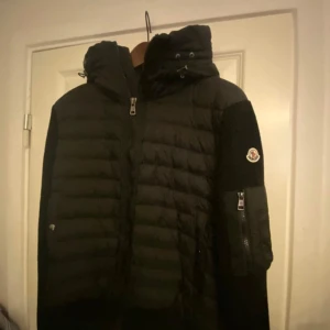 Svart dunjacka med huva - Snygg svart dunjacka från Moncler med huva och dragkedja. Jackan har en quiltad design och en praktisk ficka med dragkedja på ärmen. Perfekt för kyliga dagar.