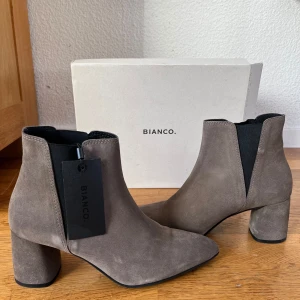 Grå mocka boots  - Snygga grå boots i mocka från Bianco med en elegant spetsig tå och bekväm klack. Perfekta för att ge din outfit en stilren touch. Elastiska paneler på sidorna för enkel på- och avtagning. HELT NYA. NYPRIS 1399. Säljer åt min mamma som inte hann lämna tillbaka dom efter att hon insett att dom var för små. Levereras med original förpackning