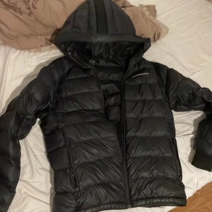 Svart dunjacka från Peak Performance - Säljer en svart dunjacka från Peak Performance. Jackan har en huva och dragkedja framtill. Perfekt för kyliga dagar med sin pufferdesign som håller värmen. En stilren och funktionell jacka för alla tillfällen.