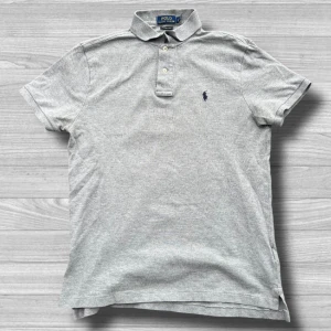 Ralph Lauren piké - Säljer en stilren grå pikétröja från Ralph Lauren i storlek M. Tröjan har en klassisk krage med knappar och det ikoniska broderade logotypen på bröstet. Perfekt för en avslappnad men ändå elegant look.