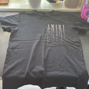 Svart t-shirt från Amiri - Säljer en svart t-shirt från Amiri med ett coolt tryck av droppande färger i flera nyanser. T-shirten har korta ärmar och en rund halsringning. Perfekt för en stilren och modern look.