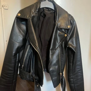 Svart skinnjacka från Zara - Snygg svart skinnjacka från Zara med dragkedjor och knappar i silver. Jackan har ett bälte i midjan och långa ärmar. Perfekt för vår/höst eller kallare sommarkväll