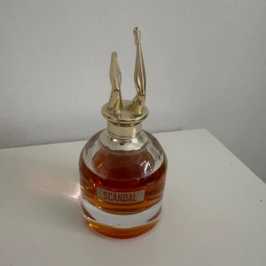 Scandal parfym - Elegant parfymflaska från Jean Paul Gaultier med namnet Scandal. Flaskan har en unik design med ett guldfärgat lock format som ben. Innehållet är en varm, bärnstensfärgad vätska som ger en lyxig känsla.
