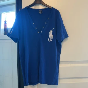 Blå t-shirt från Ralph Lauren - Säljer en blå t-shirt från Ralph Lauren med en klassisk v-ringning och knappar. Den har en broderad logga på bröstet och korta ärmar. Perfekt för en avslappnad stil.
