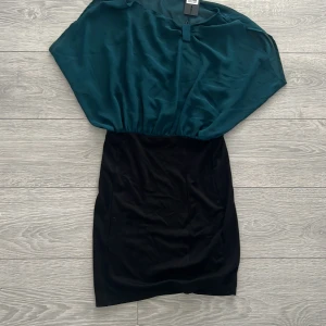 Grön och svart klänning från Vero Moda - Snygg klänning från Vero Moda med en grön överdel i ett luftigt material och en tight svart kjol. Perfekt för en stilren look. Klänningen har en elegant passform och ärmlös design.