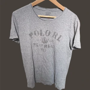 Ralph lauren T-shirt  - Säljer en grå t-shirt från Polo RL med tryck på framsidan. T-shirten har en klassisk rund hals och korta ärmar. Perfekt för en avslappnad stil.