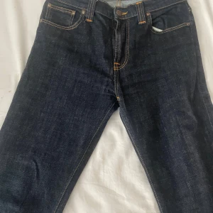 Mörkblå jeans från Nudie Jeans - Säljer ett par mörkblå jeans från Nudie Jeans i ekologisk bomull. De har en klassisk femficksdesign med orange sömmar och en knappgylf. Perfekta för dig som gillar en tidlös stil.