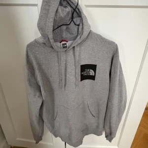 Grå hoodie från The North Face - Säljer en klassisk grå hoodie från The North Face med logga på bröstet och ryggen. Tröjan har en stor ficka framtill och en justerbar huva. Perfekt för en avslappnad stil. B-45cm L-65cm