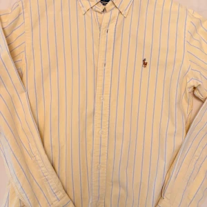 Gul och blå randig skjorta från Ralph Lauren - Säljer en stilren gul skjorta från Ralph Lauren med blå ränder och broderad logga på bröstet. Skjortan har långa ärmar och knappar framtill. Perfekt för en klassisk look.