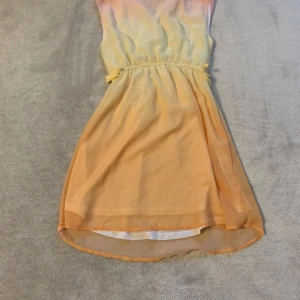 Färgglad klänning med knytband - Säljer en somrig klänning i ljusa toner av rosa, gul och orange. Klänningen har en v-ringad design och ett knytband i midjan för en smickrande passform. Perfekt för varma dagar!