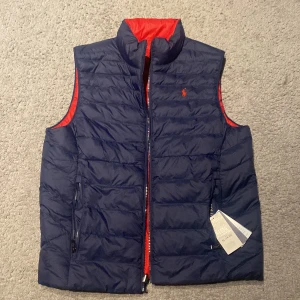 Blå och röd dunväst från Ralph Lauren - Säljer en snygg och vändbar dunväst från Ralph Lauren i blått och rött. Västen har en dragkedja framtill och är perfekt för att hålla värmen lite under våren. Den ikoniska loggan syns på bröstet. Passar perfekt för en stilren look!