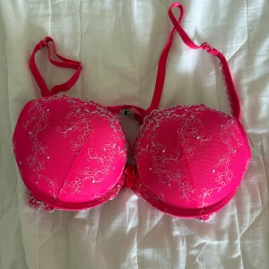 Rosa broderad pushup bh med spetsdetaljer - Snygg rosa push up bh med vackra broderade blommor och spetsdetaljer. Den har justerbara axelband och en liten rosett framtill. Perfekt för att lägga till en färgklick i garderoben. Lapp avklippt men gissar på storlek 75a eller b 