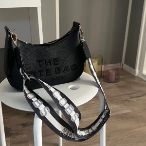 Svart väska  liknande   Marc Jacobs - Snygg svart axelväska från Marc Jacobs med texten 'THE TOTE BAG' präglad på framsidan. Väskan har en justerbar axelrem med märkets logga och en praktisk dragkedja upptill. Perfekt för att bära dina nödvändigheter med stil.