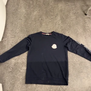 Mörkblå tröja/sweater från Moncler - Säljer en stilren mörkblå tröja från Moncler med deras logga på bröstet och text längs ärmen. Perfekt för en casual look. Tröjan har långa ärmar och en klassisk rund halsringning.